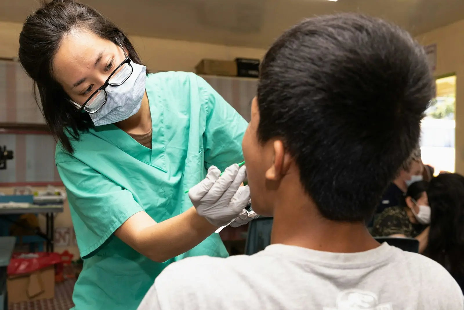 A dental practitioner in scrubs and a surgical mask treating a young patient.